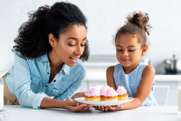 african american mother and daughter looking at homemade cupcakes in kitchen