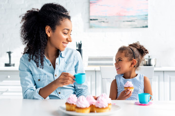 smiling african american mother and daughter with tiaras holding plastic cup and cupcake in kitchen