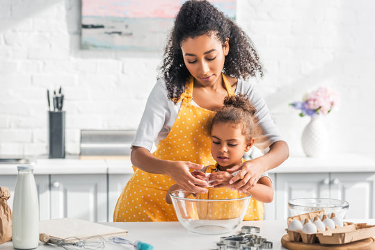african american mother helping daughter breaking egg for preparing dough in kitchen