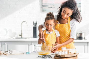 african american mother helping daughter whisking eggs for homemade dessert in kitchen