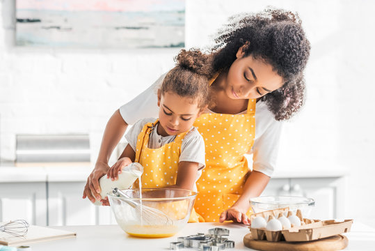 African American mother and daughter preparing dough and pouring milk into bowl in kitchen