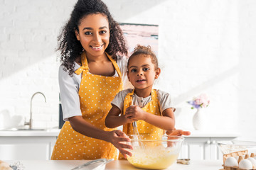smiling african american daughter and mother preparing and whisking dough in kitchen, looking at...