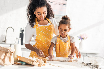 african american mother helping daughter rolling dough with rolling pin in kitchen