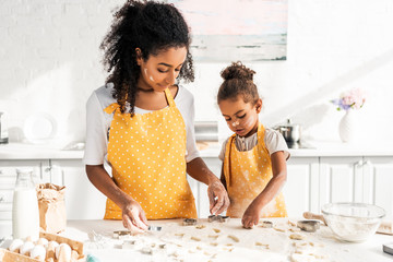african american mother and daughter in yellow aprons preparing cookies with molds in kitchen