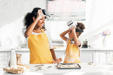 african american mother and daughter preparing dough in virtual reality headsets and looking at each other in kitchen