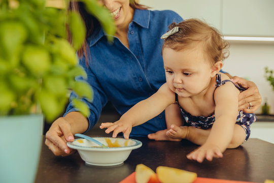 Mother with baby daughter eating fruit pulp in kitchen