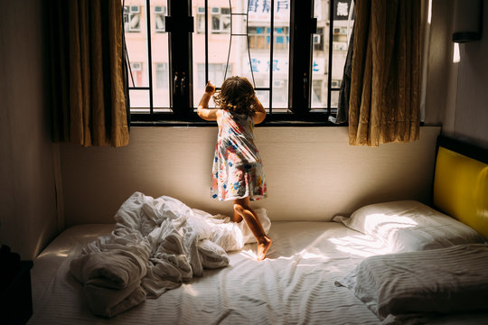 China, Hong Kong, Mong Kok, back view of little girl standing barefoot on bed looking out of window