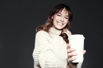 Beautiful woman wearing sweater shows paper disposable coffee cup. Drinking coffee, smiling and laughing.