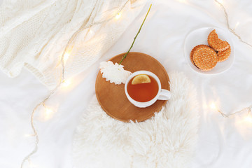 Cup of tea, white flower, wooden tray, cookies, fluffy plaid, knitted sweater and christmas garland lights on white bedding at home. Breakfast in bed. Cozy winter homely scene. Flat lay, top view.