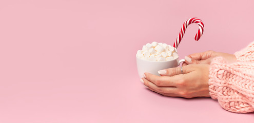 Female hands in knitted sweater holding cup of marshmallows and Christmas candy cane on pink background Flat Lay copy space. Winter traditional food Festive decor celebration presents Xmas holiday