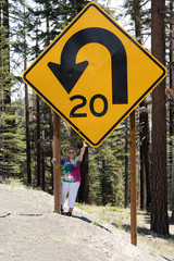 An adult woman stands next to a giant road sign in Mammoth Lakes, California, showing the scale of the size of the sign. Giving peace sign