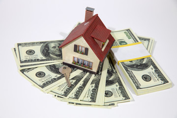 A small house lies on a fan of hundred dollar bills. The keys to the purchased house. Reduced copy of the house on a white background.