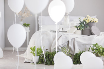 Flowers and green plants on bed with blanket in grey bedroom interior with white balloons. Real photo
