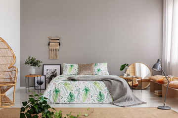Real photo of double bed with leafy sheets placed in bright bedroom interior with macrame on the wall, mirror and posters on the floor and fresh plants