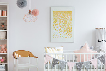 Stylish white and brown chair next to crib with pillows in trendy baby room with golden poster on...