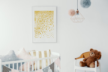 Abstract golden painting in white frame above wooden crib with pillows and chair with teddy bear in scandinavian baby room interior - obrazy, fototapety, plakaty