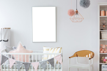 Stylish white and brown chair next to crib with pillows in trendy baby room with empty mockup poster on the wall