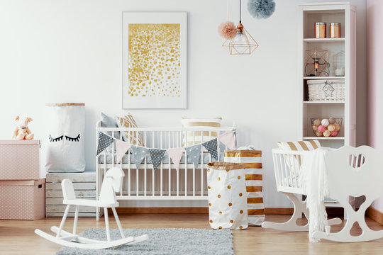 Wooden rocking horse, white and gold paper bags and white wooden crib in cozy baby nursery with painting on the wall