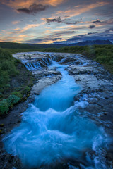 The waterfall Bruarfoss with golden clouds in the sky. The flowing water is captured by a long exposure. Amazing blue color of water from the glacier. Natural and colorful environment.
