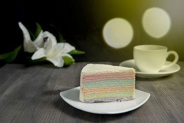 Rainbow crape cake on white plate with a cup of hot tea on dark wooden table, isolated black background and bokeh light.