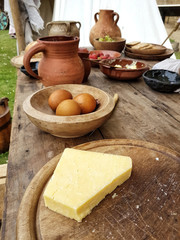 A lump of white cheese on a board, with medieval pots and bowls of food  arranged behind it.