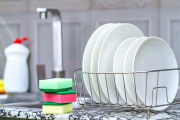 Different clean plates in dish drying rack, dish sponges and dishwashing detergent on the table on...
