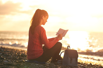 A young, happy woman listening to music and writes her thoughts in a personal diary on the beach at sunset. Enjoy your time and moments. Travel and relaxation