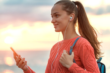 A young, happy woman listening to music and enjoys beautiful seascape on the beach at sunset. Travel and relaxation