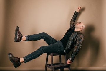 Indoor portrait of artistic adult unusual male. Funny bald man in biker leather jacket and oxford shoes. Fashion clothes. Stylish weird boy falling down from chair on brown background. Movie scene. © benevolente