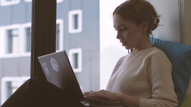 Beautiful girl at the window working on a laptop. Against the background of the facade of the business center. Corresponding with colleagues after lunch break.