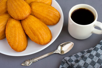 Delicious madeleine, homemade traditional french pastry (small cookie) on grey tablecloth. Cup of coffee, spoon, grey napkin.