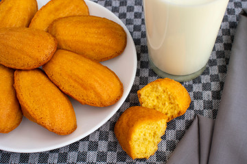 Delicious madeleine, homemade traditional french pastry (small cookie) on grey tablecloth. Glass of milk, halves of madeleine, grey napkin.