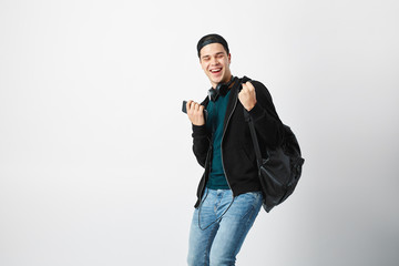 Fototapeta na wymiar Happy guy with a black backpack on his shoulder and headphones on his head dressed in a dark t-shirt, jeans, sweatshirt and a cap uses mobile phone in the studio on a white background