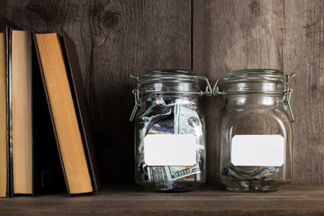Two piggy banks, with different amounts of money and space for text, stand on a wooden bookshelf.