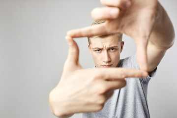 Plakat Blond guy dressed in a white t-shirt keeps his hands in front of him making with fingers a shape of a frame on the white background in the studio