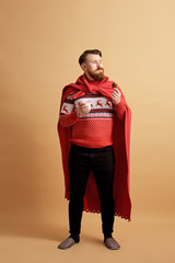 Red-haired man with beard dressed in a red and white sweater with deer and red cape  fluttering in the wind holds a glass of milk and cookie on a beige background