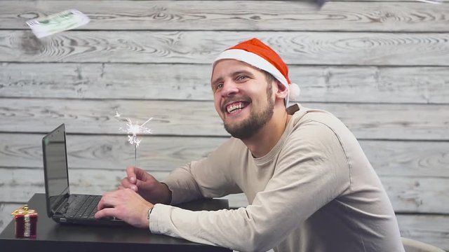 dreamy man in a christmas hat with bengal fire sparkler and laptop in his hands. Falling Money Dollars.