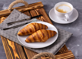 Croissant, a cup of coffee at wooden tray with napkin on a gray stone concrete table.