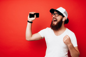 Portrait of happy bearded hipster man singing and listening to music with cell phone and wireless earphones isolated over red wall