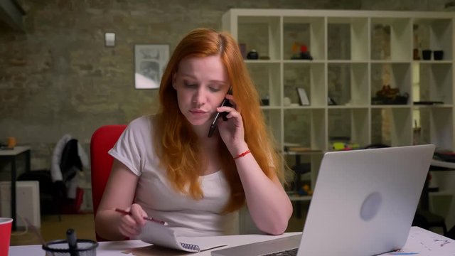 Hard-working ginger caucasian businesswoman is talking over phone and checking nites near laptop, office illustration, pretty person