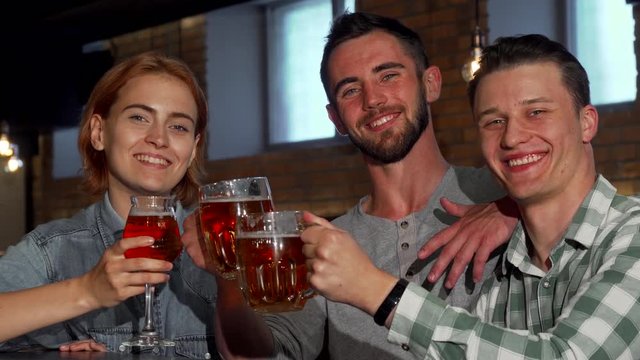 Happy young people clinking their beers and smiling to the camera. Group of friends enjoying drinking beer together at the pub. Cheerful woman and her male friends partying. Leisure concept.