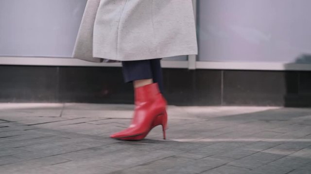 Legs of unrecognizable woman wearing black pants, white coat and red boots walking in autumn street. Tracking slow motion medium shot