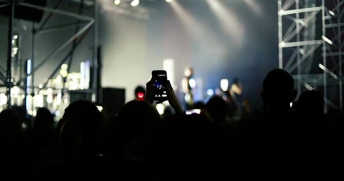 Silhouettes of people enjoying the concert of favorite singer. They waving hands and giving him applause the song. Fans dancing and shooting mobile videos at the concert in crowded music hall