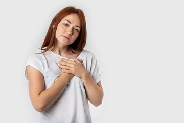 Portrait of a red-haired young woman keeps hands on chest, expresses sympathy. Kind hearted friendly nice girl shows kindness, wearing blank white t-shirt