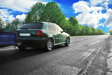 Fototapeta premium A green car with a trailer on the countryside road against sky with clouds