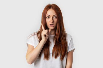 Beautiful redhead woman keeping finger pointed upwards, showing something above her head, making gesture with index finger.