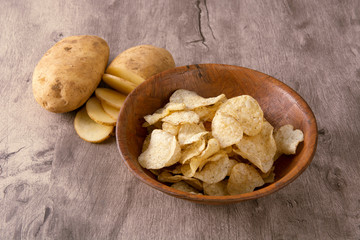 Potato chips on a bowl and raw sliced potato over wood background