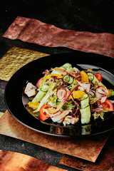 salad with ham, orange slices, tomato, cucumber and lettuce leaves in a black plate on a copper background. close up