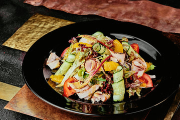 salad with ham, orange slices, tomato, cucumber and lettuce leaves in a black plate on a copper background. close up
