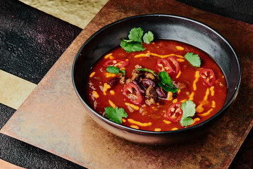spicy tomato soup with beans, chili pepper, cheese and parsley in a black plate on a copper background. close-up.